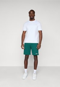 Hvid t-shirt med kort ærme parret med grønne shorts med et logo. Modellen bærer hvide sneakers og ankelsokker. Simpelt, atletisk design.