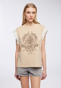 T-shirt in cotone beige con maniche frangiate, caratterizzata da un motivo grafico centrale marrone a fiori e dal testo "VAI CON IL FLUSSO."