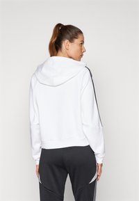 White cropped hoodie with a hood, featuring black side stripes. The material appears soft, with a relaxed fit and ribbed hem.