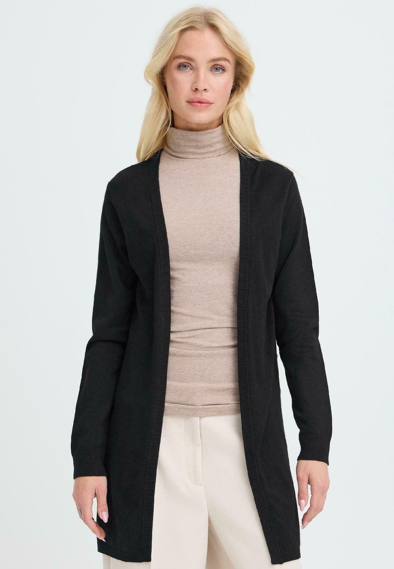 Črn cardigan z dolgim, odprtim dizajnom in rebrastimi manšeti, oblečen čez bež turtleneck. Gladka tekstura, minimalne podrobnosti.