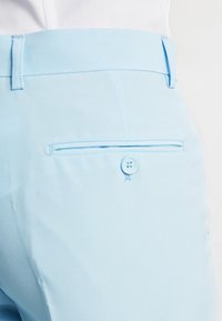 Světle modré kalhoty na míru s klopou zadní kapsy na knoflík a poutky na opasek, nošené s bílou košilí zasunutou dovnitř.