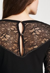 Zwarte top met een kanten achterkant met bloemmotieven. Sleutelgatdetail bij de hals, bevestigd met een klein knoopje. Glad stoffen basis en afwerking.