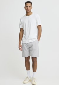 T-shirt blanc en coton avec manches retroussées, short gris à cordon de serrage, et baskets crème à rayures. Le mannequin pose sur un fond clair.