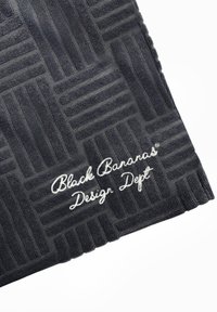 Zwarte handdoek met een gestructureerd, gemarmerd oppervlak in donkergrijs. Bevat witte geborduurde tekst: "Black Bananas® Design Dept." aan een rand.