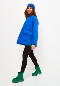 Kék steppelt dzseki, elülső zsebbel, zöld felső és fekete leggings felett viselve. Zöld bokacsizmák vastag talppal.