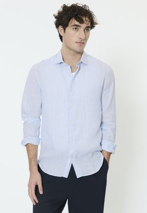 Mees lokkis juustega, kes kannab helesinise ja valge triibulise nööbitava särgi käised üles rullitud ja tumedaid pükse, seisab valge tausta ees.