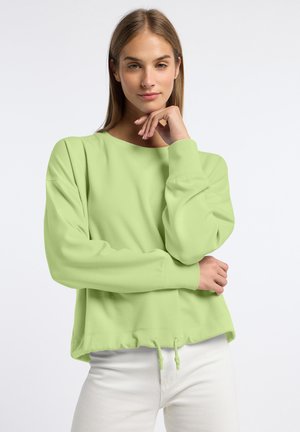 Jonge vrouw met lichtbruin haar, gekleed in een lichtgroene lange-mouw sweater en witte broek, poseert voor een effen witte achtergrond.