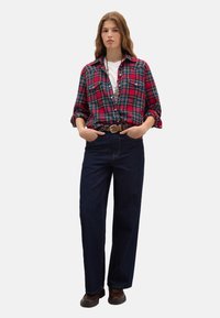 Red and green plaid flannel shirt layered over a white t-shirt, paired with dark blue wide-leg jeans and brown boots.