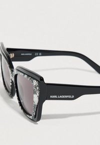 Μαύρα γυαλιά ηλίου με τετράγωνο σχήμα και υφή σε λευκό και γκρι μοτίβο στα πλαίσια. Οι φακοί είναι σκούροι και στην πλευρά φέρουν το όνομα της μάρκας "KARL LAGERFELD".