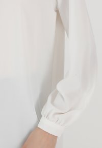 Weiße Bluse mit langen, aufgeplusterten Ärmeln und einem gesammelten Bündchen. Der Stoff ist glatt und leicht, mit einem klaren, minimalistischen Design.