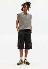 Grijs mouwloos shirt, donkerdenim cargo shorts met zijzakken en olijfgroene sneakers met gele accenten. Model staat tegen een neutrale achtergrond.