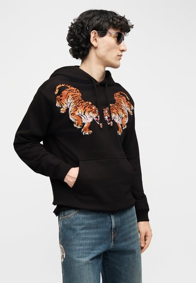 Jeune homme portant un sweat à capuche noir avec deux tigres brodés sur la poitrine, un jean bleu et des lunettes de soleil, posant de profil avec la main dans la poche.