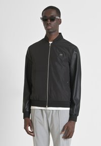 Black bomber jacket with a zip front, fabric mixed with sleek leather sleeves, ribbed cuffs and trim, featuring a small logo patch on the chest.