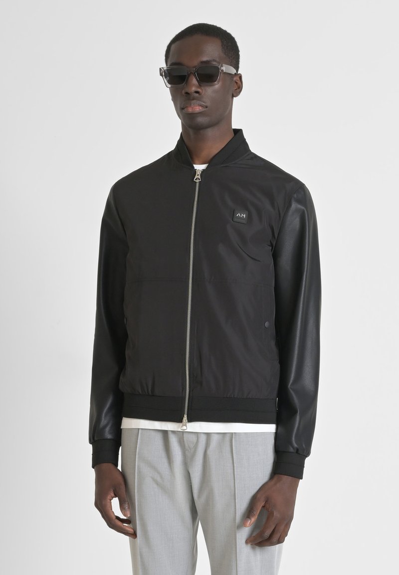 Black bomber jacket with a zip front, fabric mixed with sleek leather sleeves, ribbed cuffs and trim, featuring a small logo patch on the chest.