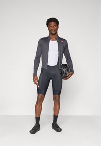 Tenue de cyclisme : veste grise ajustée à manches longues, couche de base en maille blanche, cuissards rembourrés noirs, chaussettes noires et chaussures de cyclisme. Tenant un casque noir.