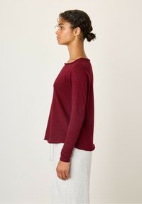 Burgundy langærmet top med rund halsudskæring og rå kantdetaljer. Stof ser blødt ud; modellen har den på over lysegrå bukser.