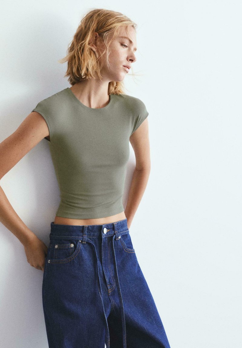 Femme aux cheveux blonds courts portant un crop top ajusté vert olive et un jean large taille haute bleu foncé, se tenant contre un mur pâle.