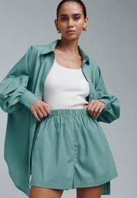Padidinta žalsvai mėtų spalvos marškiniai su pūstomis rankovėmis, derinami su baltu ribuotu tank topu ir atitinkančiais šortais su elastine juosmens juosta.