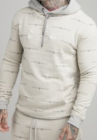 Sudadera beige con capucha gris, con un logo bordado y un patrón de texto repetido. Elaborada en una mezcla de algodón y con bolsillo delantero.