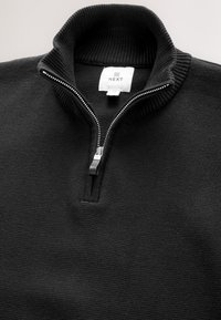 Pull-over noir en maille côtelée avec une fermeture éclair argentée partiellement zippée et une étiquette de marque "NEXT" à l'intérieur du col.