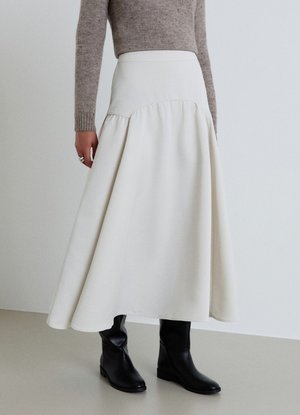 Donna che indossa una gonna midi a vita alta color crema con orlo svasato, abbinata a un maglione beige e stivali neri a metà polpaccio.