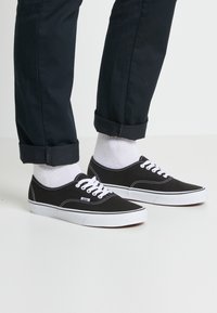Eine Person trägt hochgerollte schwarze Hosen, weiße Socken und schwarze Sneakers mit niedrigerem Schnitt, die weiße Sohlen und Schnürsenkel haben, und steht auf einer weißen Fläche.