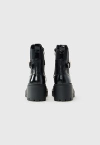 Bottines noires à talons hauts avec semelles épaisses, fermetures éclair latérales et boucles décoratives, présentées de dos sur un fond blanc.