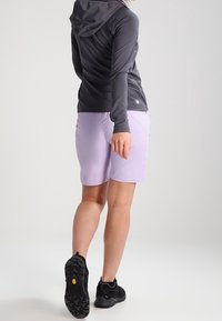 Persona vestida con una chaqueta con capucha negra, pantalones cortos morados claros y zapatos de senderismo negros, de pie con la espalda hacia la cámara.