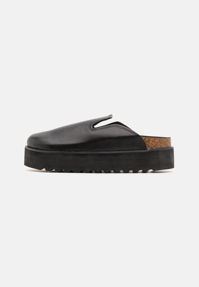 Svart läder slip-on sko med en kraftig svart gummisula, texturerad korkklack och utskärningar på sidorna.