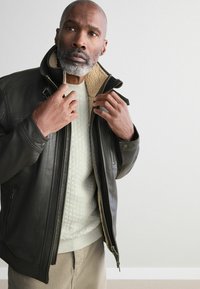 Chaqueta de piel negra con un cuello de piel sintética, bolsillos con cremallera y un suéter beige acolchado debajo. El modelo ajusta la chaqueta.