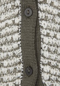 Stickad cardigan med en texturerad vit och grå mönster, med en enfärgad olivgrön knappslå och tre svarta knappar i mitten.