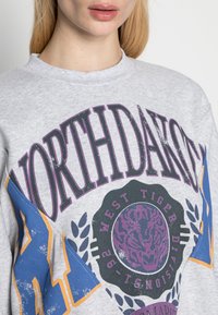 Šedá mikina s výrazným grafickým textem "NORTHDAKOTA" v purpurové barvě, s modrými akcenty a kruhovým emblémem "WEST TIGER" na přední straně.