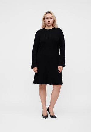 Femme aux cheveux blonds portant une robe noire à manches longues et des talons hauts noirs, debout devant un fond clair uni.