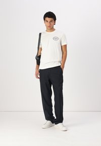 White cotton t-shirt with black oval logo, paired with dark trousers and white sneakers. Simple design with a relaxed fit.