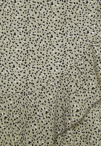 Tissu vert olive avec un motif tacheté noir et blanc irrégulier et un détail volanté diagonal.