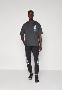 Graues Baumwoll-T-Shirt mit blauem grafischem Design auf der Vorderseite, kombiniert mit schwarzen Jogginghosen mit weißen Streifen an den Seiten. Schwarze Sneaker.