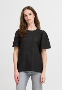 Blusa nera texture con maniche a sbuffo corte, scollatura rotonda e vestibilità leggermente ampia. Indossata con jeans in denim grigio chiaro.