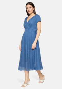 Femme portant une robe bleue scintillante jusqu'aux genoux, à manches courtes et décolleté en V profond, associée à des sandales blanches compensées.