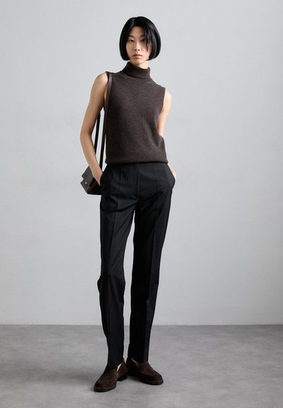 Rjav brezrokavnik s turtleneck ovratnikom, krojene črne hlače in temno rjavi semiš loaferji. Roke v žepih, vidna preprosta usnjena torba.