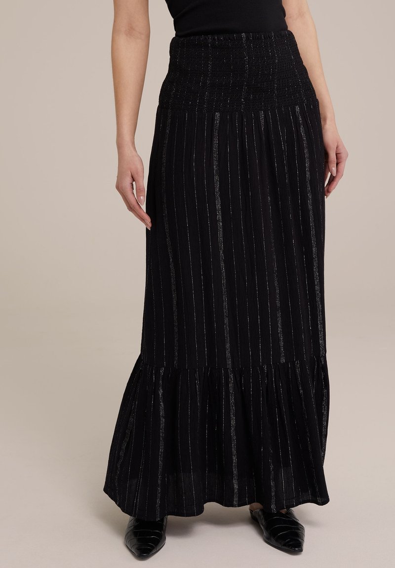 Femme portant une longue jupe noire avec de fines rayures verticales argentées, une ceinture froncée et un ourlet volanté, associée à des chaussures noires pointues.