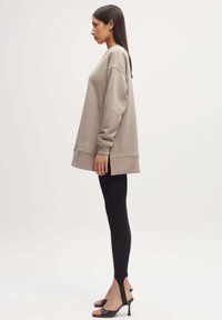 Overmaat beige sweatshirt met geribbelde manchetten, zijsplitten en een subtiele logo aan de zoom, gecombineerd met zwarte leggings en sandaals met hak.