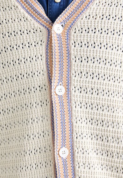 Strickjacke in Creme mit Lochmuster. Verfügt über eine gepunktete Leiste in Orange und Blau sowie weiße Kunststoffknöpfe.
