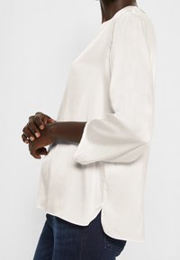 Weiße Bluse mit langen Ärmeln und glatter Satin-Oberfläche, mit einem runden Ausschnitt und seitlichen Schlitzen. Getragen mit dunkelblauen Jeans.