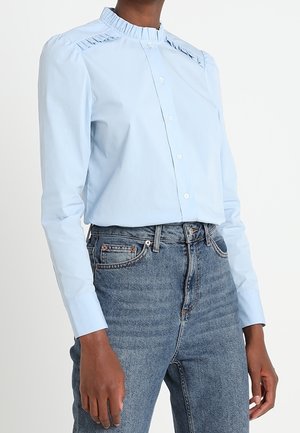 Light blue long-sleeve blouse with pleated collar and shoulder details paired with high-waisted blue denim jeans.