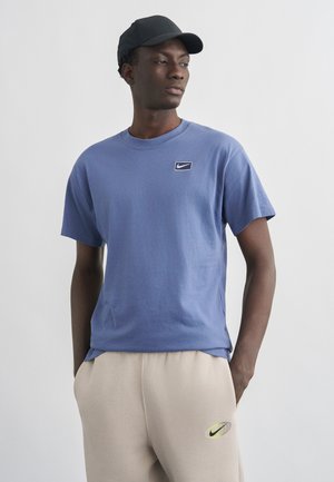 Egy férfi kék Nike pólóban, bézs Nike melegítőnadrágban és fekete sapkában, zsebre tett kézzel áll, oldalra nézve.