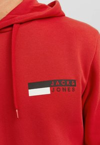 Sudadera roja con capucha ajustable. Presenta un logo impreso en blanco y negro: "JACK & JONES" en la parte delantera. Material de algodón suave.