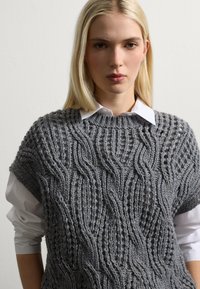 Jonge vrouw met steil blond haar, gekleed in een wit shirt onder een grijze gebreide vest met textuur, staande tegen een effen lichte achtergrond.