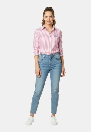 Giovane donna in piedi, indossa una maglietta a maniche lunghe rosa e bianca a righe, jeans blu e sneakers bianche, con i capelli legati indietro.
