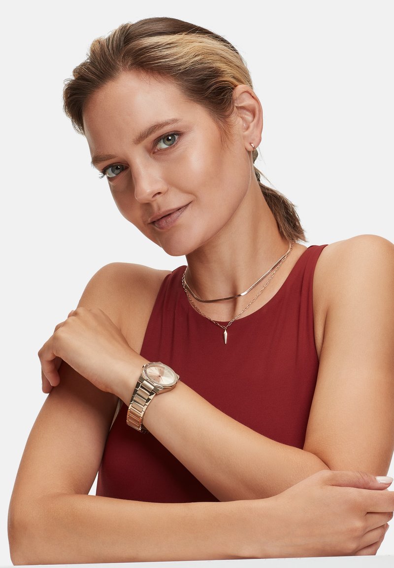 Goudkleurig polshorloge met een ronde wijzerplaat, metalen band en een lichtroze klok. Gelaagde zilveren kettingen en een bordeaux top maken de look compleet.