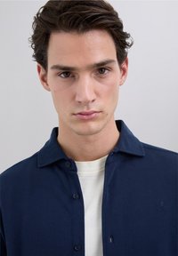 Camisa azul marino con cuello y cierre de botones, tela texturizada, usada sobre una camiseta blanca; presenta un logo bordado en el pecho.